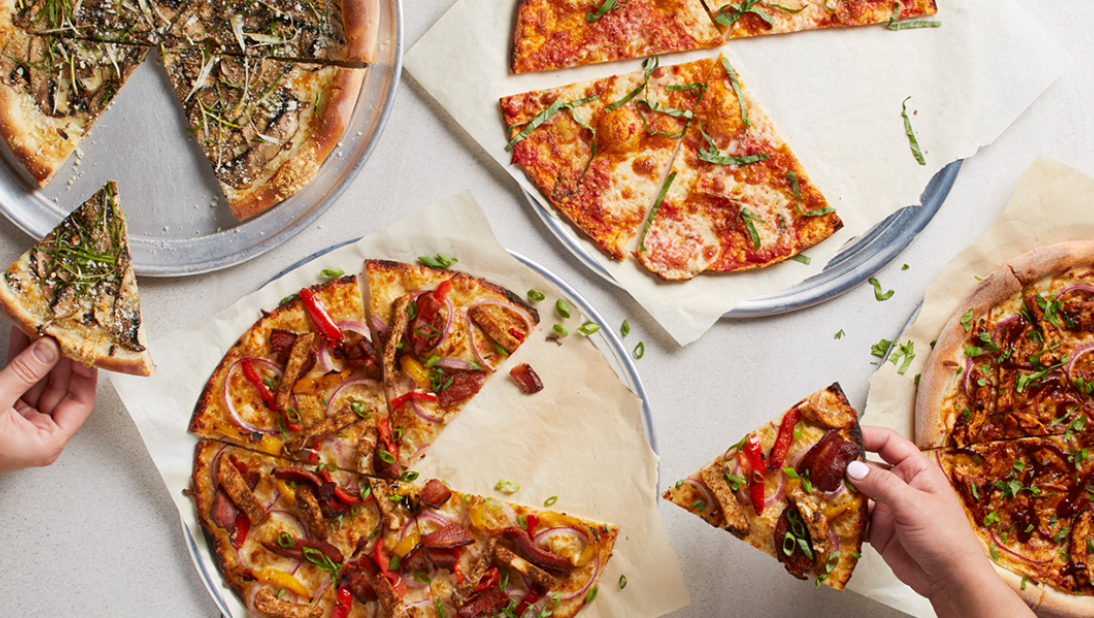 Four pizzas with various toppings, some partially sliced, are arranged on a table—just like you’d find at popular pizza chains. Two hands reach in to pick up a slice each, suggesting a casual meal shared among friends.