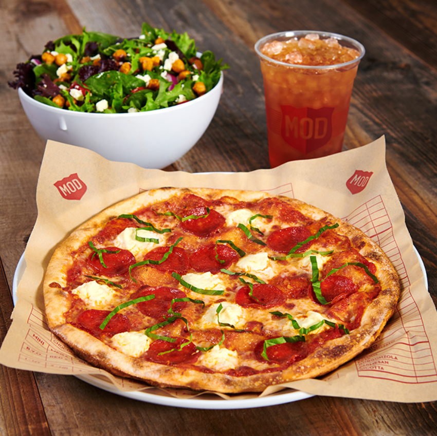 A thin-crust pepperoni pizza with dollops of ricotta cheese and fresh basil, reminiscent of what top pizza chains serve, sits on a plate beside a mixed greens salad with cheese and a cold iced tea in a plastic cup on a wooden table.