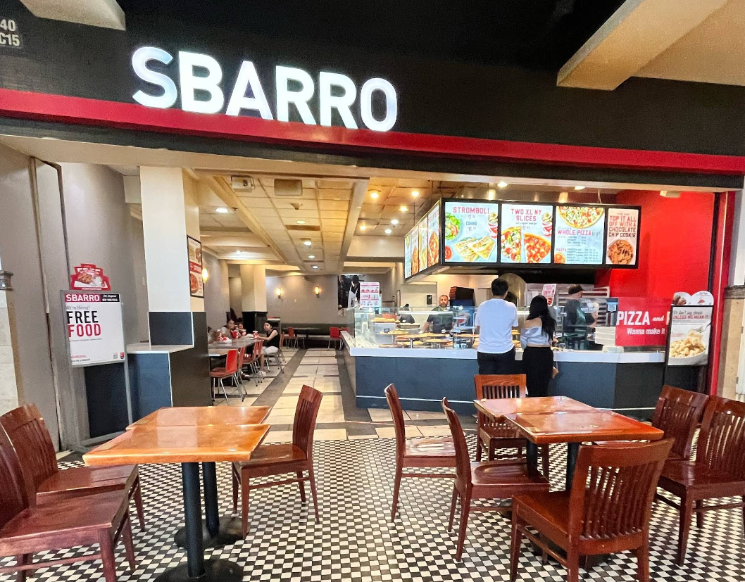 A Sbarro restaurant, part of popular pizza chains, features a counter with menu boards, two people ordering, empty tables and chairs in the foreground, and a sign on the left offering free food.