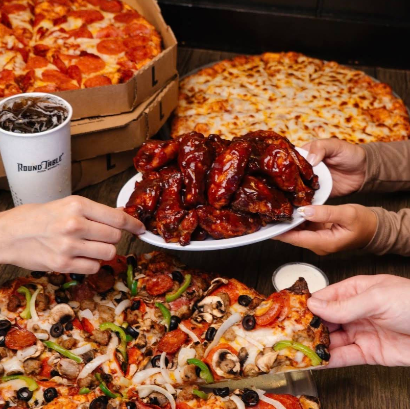 Three people share pepperoni and veggie pizzas from popular pizza chains, saucy chicken wings, a cup of soda, and ranch dip on a wooden table. Pizza boxes are open in the background.