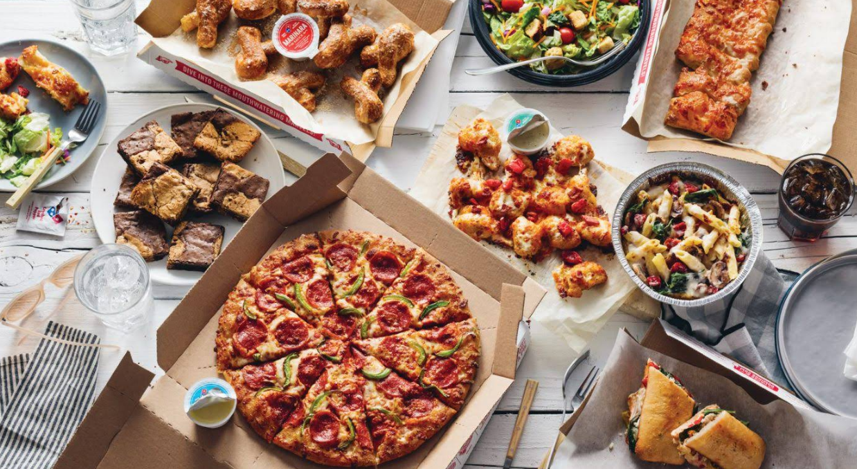 A table filled with various foods from popular pizza chains, including pizza, breadsticks, brownies, salad, chicken bites, pasta, a sandwich, and soft drinks in takeout containers.