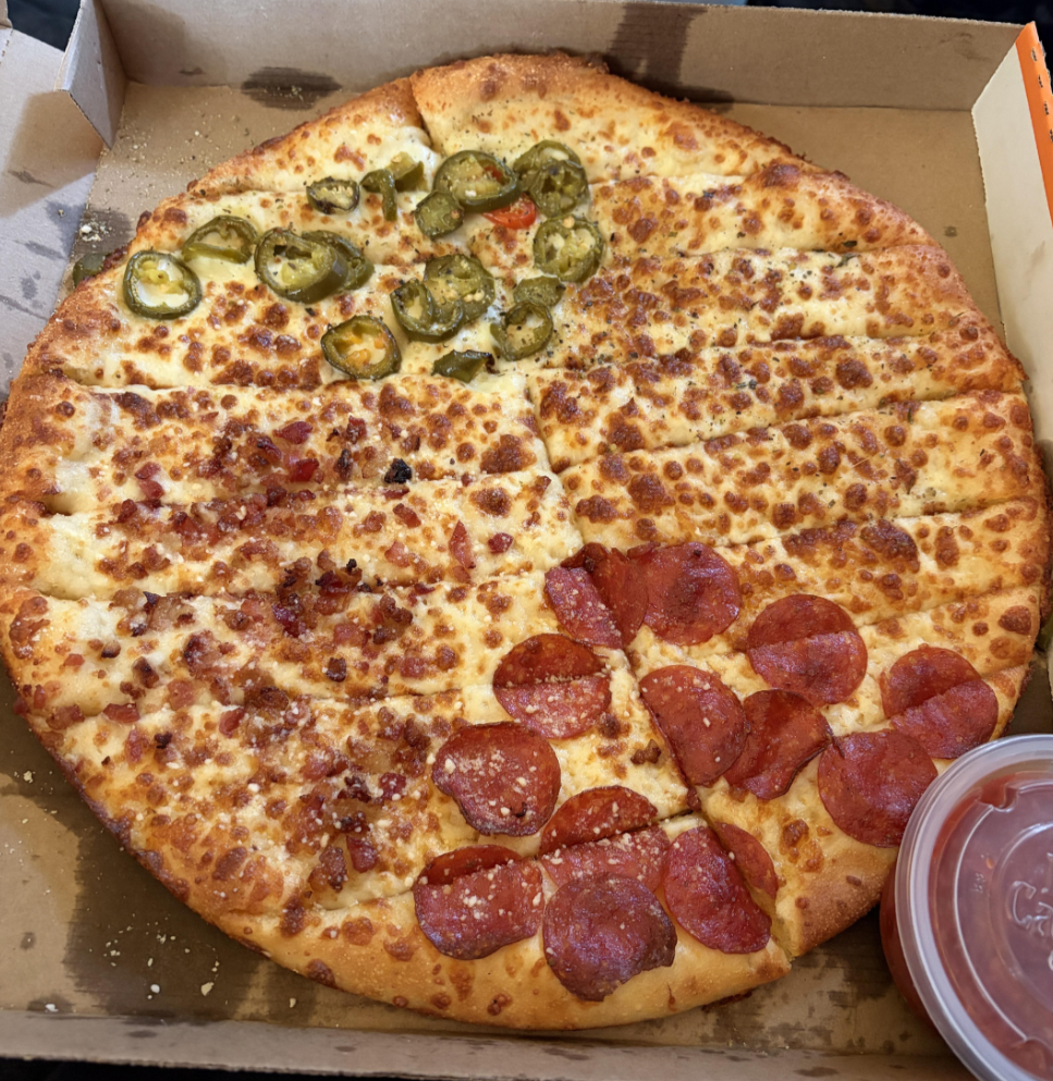 A pizza, served in the style of popular pizza chains, is cut into strips with three topping sections: jalapeños on cheese, bacon bits on cheese, and pepperoni. A cup of sauce sits in the bottom right corner of the open pizza box.