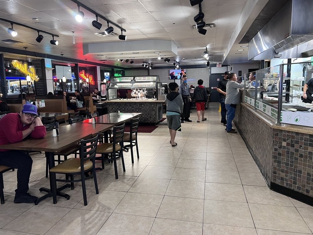 A spacious restaurant interior with several diners; booths are on the left, tables are in the center, and a buffet area—famous among breakfast burrito spots—is on the right. Some people walk or stand, while one person sits alone at a table.