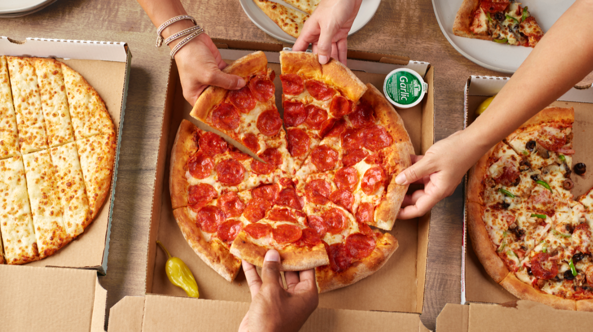 Three people grab slices from open boxes of pepperoni, cheese, and veggie pizzas—classic offerings from popular pizza chains. A cup of garlic sauce and a pepperoncini sit beside the pepperoni pizza.