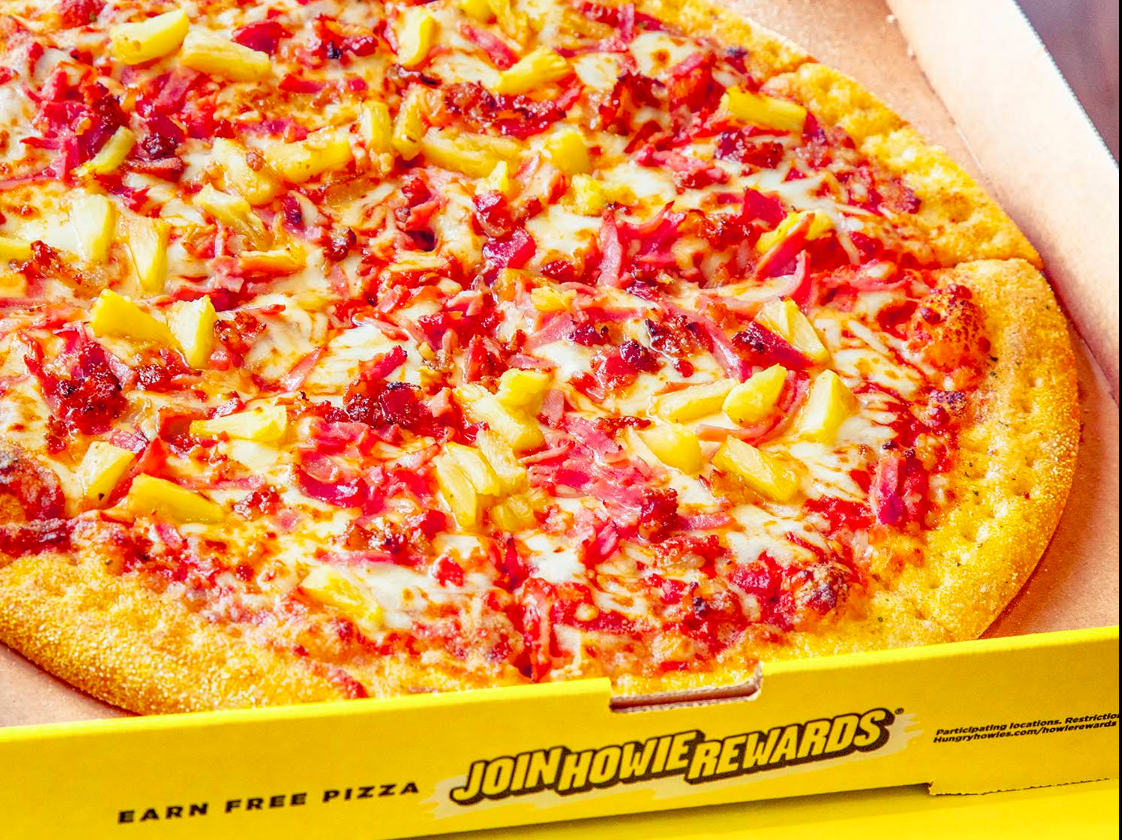 A Hawaiian pizza from one of the top pizza chains, topped with ham, bacon, pineapple, and melted cheese, sits in an open box with “JOIN HOWIE REWARDS” on the yellow edge.