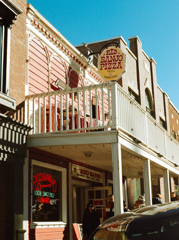 A red and white building with a sign reading "Red Banjo Pizza Pasta Sandwiches," a neon window sign, and people entering the restaurant—a local spot that stands out from big pizza chains under the clear, blue sky.