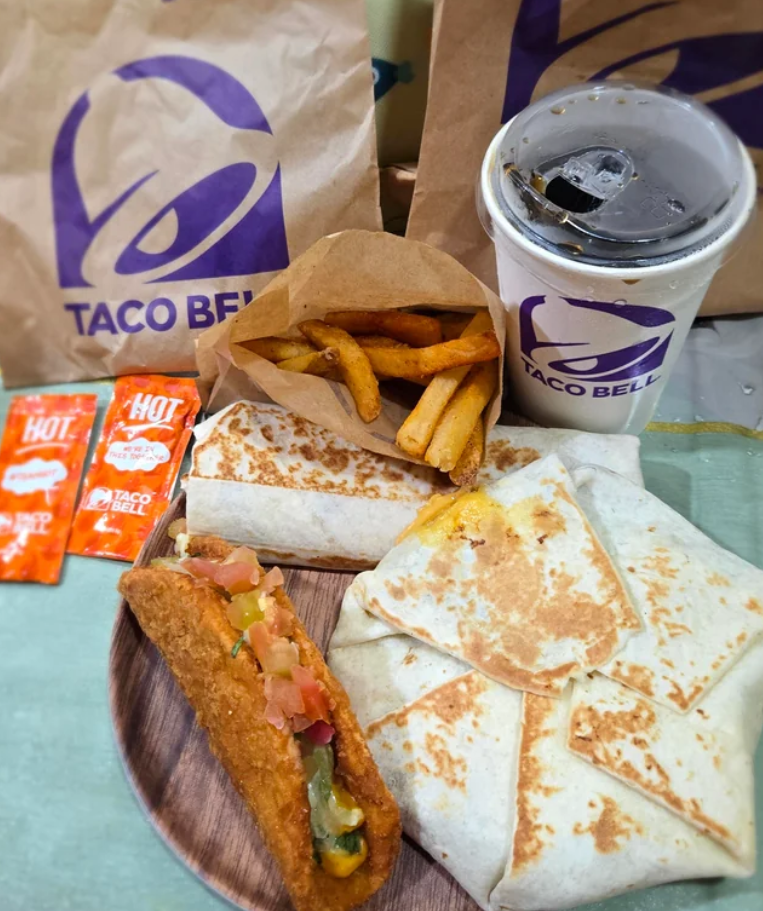 A Taco Bell meal with a quesadilla, Crunchwrap, fries, chicken taco, hot sauce packets, and a drink on a tray—classic fare from one of the most popular Mexican food chains. Taco Bell-branded bags and cup are in the background.