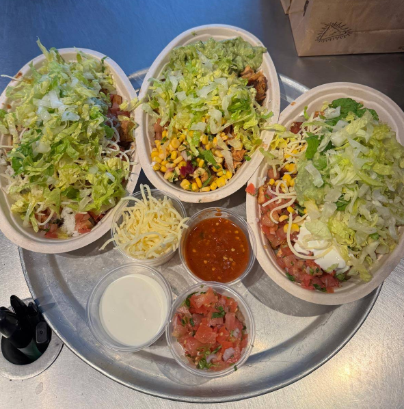 Three burrito bowls topped with shredded lettuce, cheese, corn, and more are served on a metal tray with sides of shredded cheese, salsa, sour cream, and pico de gallo—classic flavors you’ll find at popular Mexican food chains.