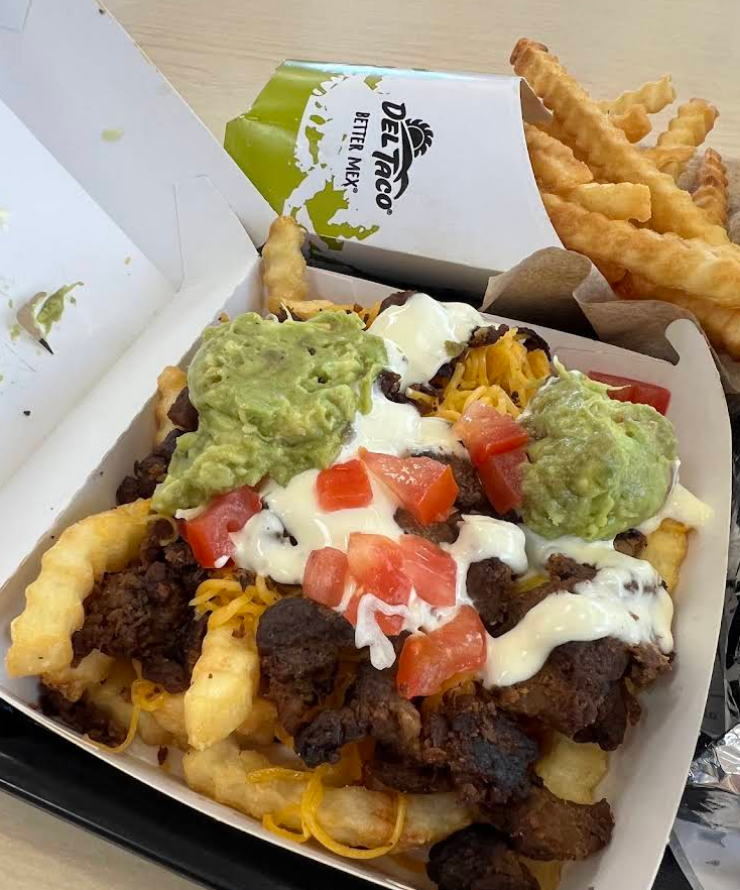 A box of crinkle-cut fries topped with grilled steak, melted cheese, diced tomatoes, guacamole, and creamy sauce—a flavorful twist typical of top Mexican food chains. In the background, a side of plain crinkle-cut fries sits in a Del Taco container.