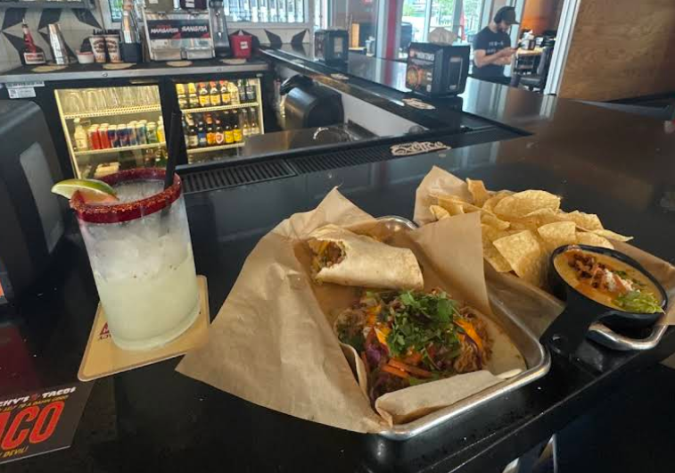 A margarita, a tray with a burrito and taco, tortilla chips, and a bowl of queso are set on a bar counter inside a casual restaurant, capturing the lively vibe often found in popular Mexican food chains. Drink coolers and bar supplies are visible in the background.