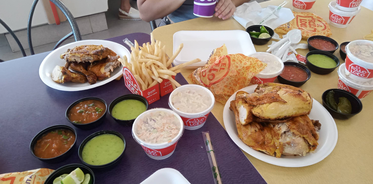 A table with plates of grilled chicken, French fries, coleslaw, sauces, lime wedges, and El Pollo Loco packaging from one of the top Mexican food chains. A person is seated at the table holding a cup.