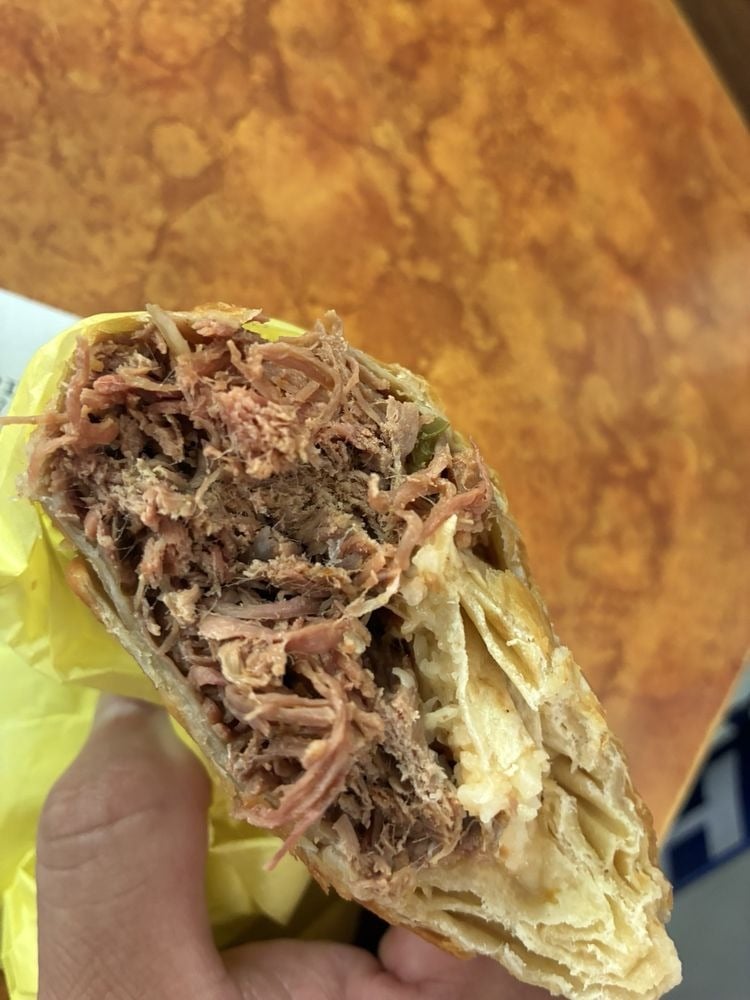 A close-up of a hand holding a half-eaten burrito filled with shredded beef, wrapped in yellow paper—perfect inspiration for breakfast burrito spots—set against a brown, marbled tabletop in the background.