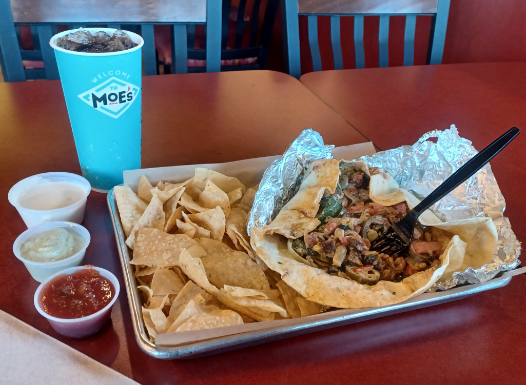 A tray from one of the popular Mexican food chains features tortilla chips, three sauces in small containers, and a foil-wrapped burrito bowl with a black fork. A large blue Moe’s drink cup sits behind it on a red table with chairs in the background.
