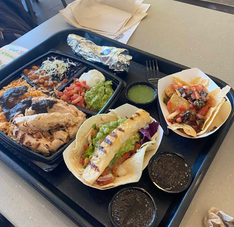 A black tray holds grilled chicken with rice and beans, a foil-wrapped burrito, two tacos with grilled fish and vegetables, tortilla chips with salsa, and containers of green and dark salsa—classic flavors found at top Mexican food chains.
