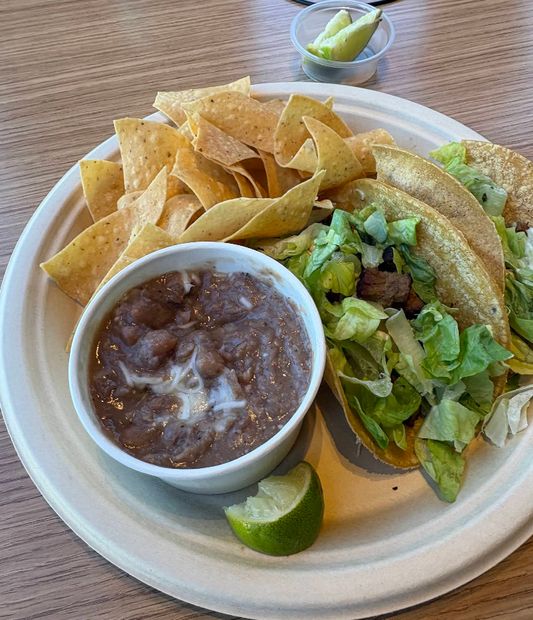 A plate with tortilla chips, a cup of refried beans, a lime wedge, and a taco filled with lettuce and meat—just like you’d find at popular Mexican food chains. In the background, sliced jalapenos and lime sit on a wooden table.