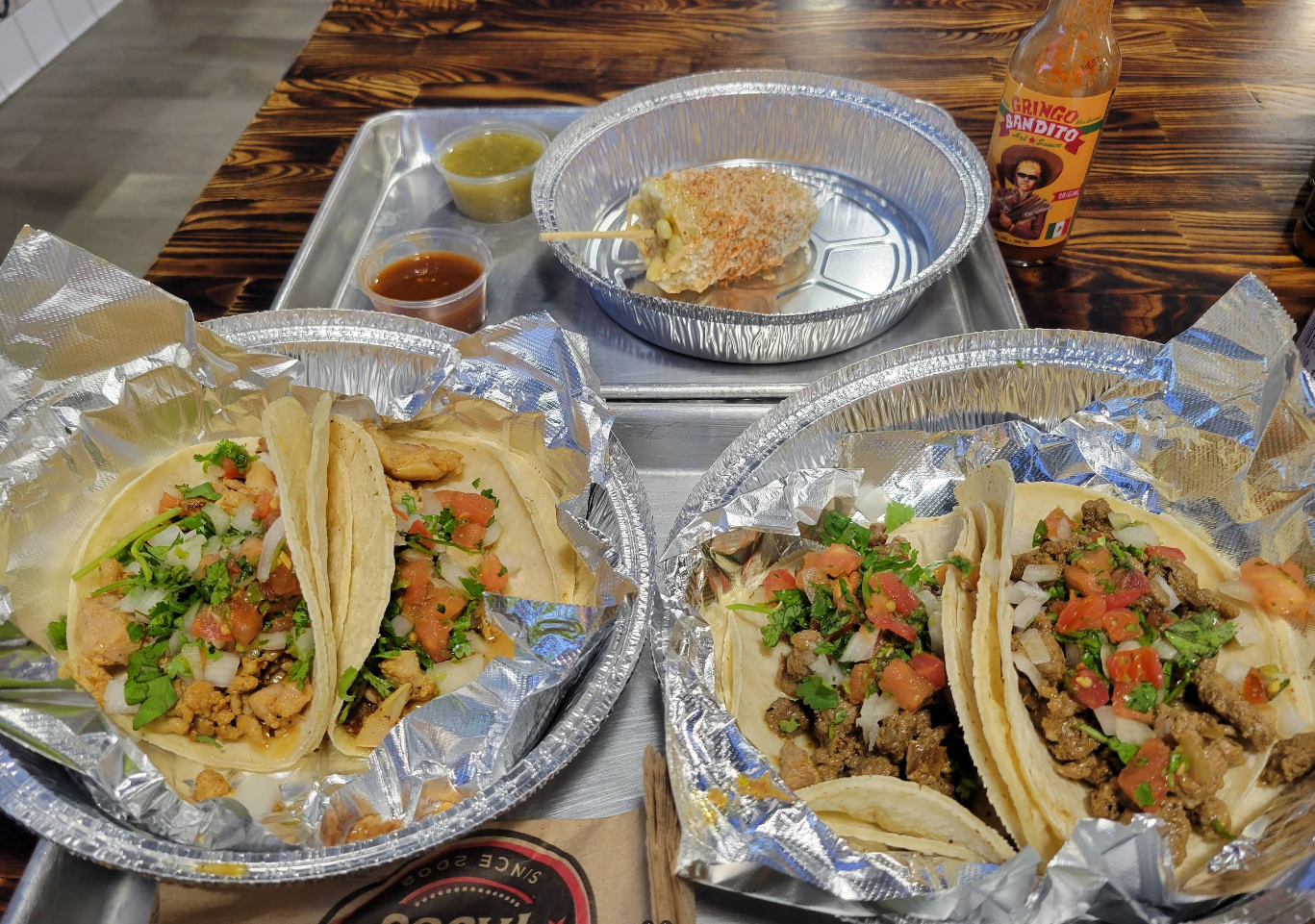 Two foil trays with soft shell tacos topped with chopped onions, cilantro, and tomatoes, plus a grilled corn cob, three salsa containers, and a bottle of hot sauce—just like you’d find at top Mexican food chains—on a wooden table.