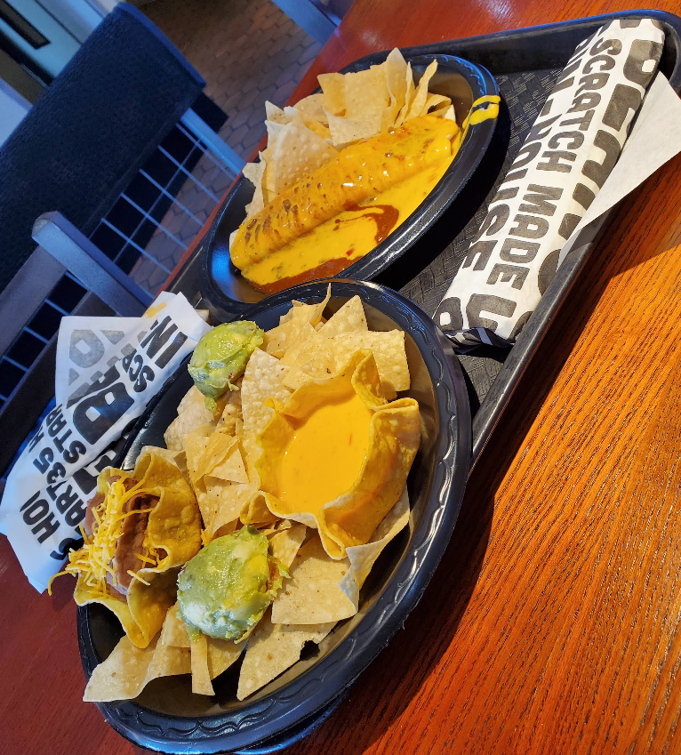 A tray with two black bowls: one holds tortilla chips with cheese dip and guacamole, the other has a large burrito topped with cheese sauce—classic offerings you’d find at popular Mexican food chains, all served on a wooden table in a restaurant setting.