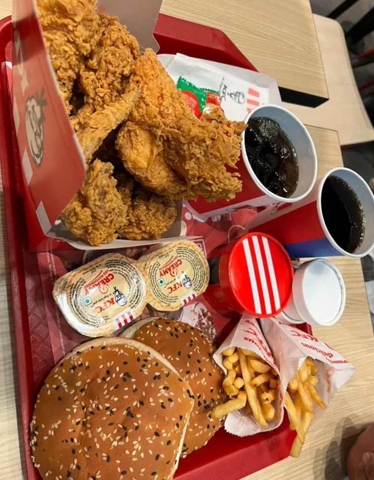 A red tray with fried chicken pieces from popular fried chicken chains, two burgers with sesame seed buns, two packs of fries, three drink cups, and packets of butter, on a light wooden table.