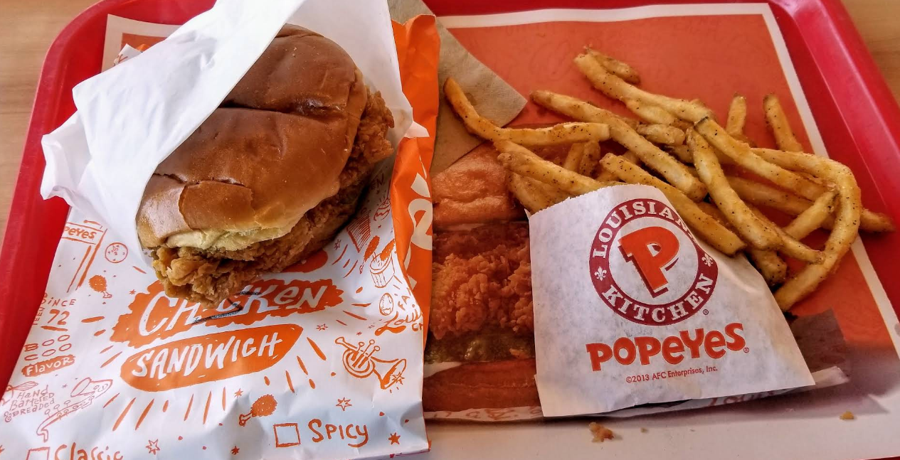 A fried chicken sandwich partially wrapped in orange and white paper sits on a tray next to seasoned French fries in a Popeyes-branded pouch, showcasing why Popeyes is a standout among fried chicken chains.