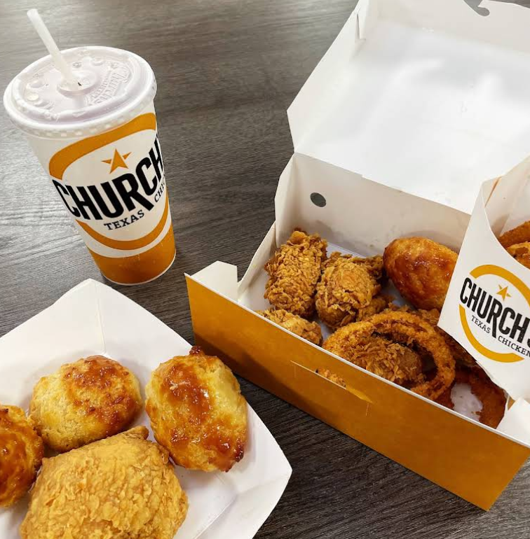 A meal from one of the top fried chicken chains, Church’s Texas Chicken, featuring a drink, several pieces of fried chicken, onion rings, and three golden biscuits on a wooden table.