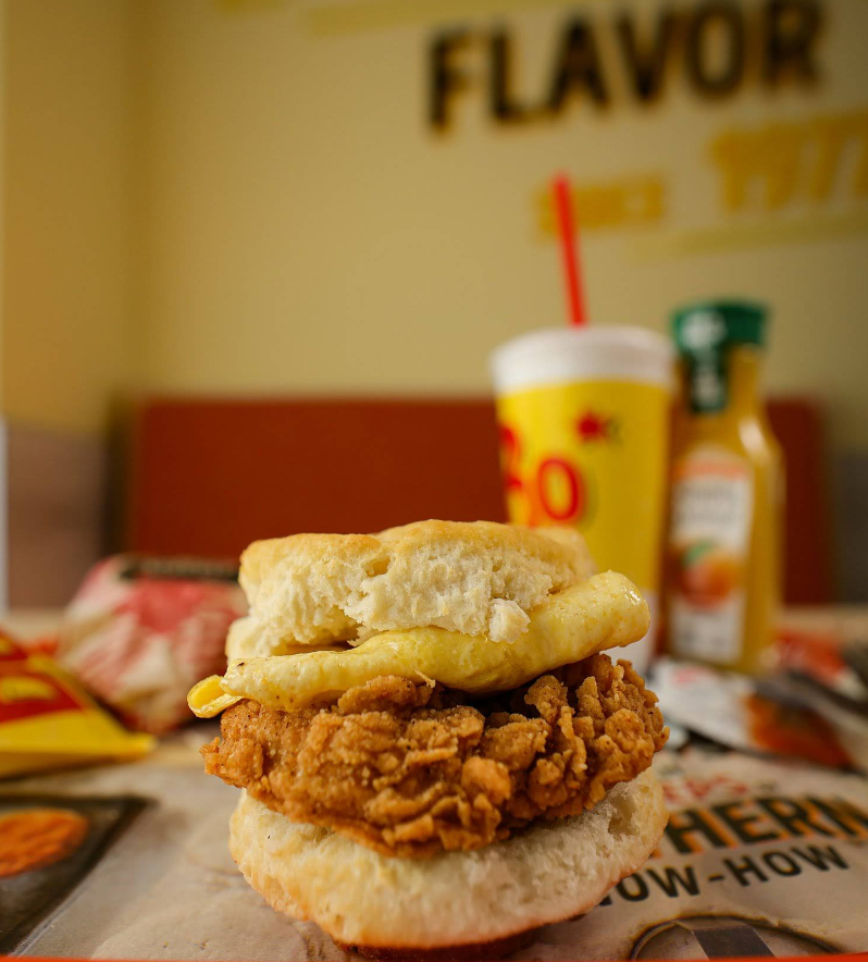 A close-up of a breakfast sandwich with fried chicken, egg, and biscuit on a table at a fast-food restaurant—reminiscent of popular fried chicken chains—with a drink cup and juice bottle blurred in the background.