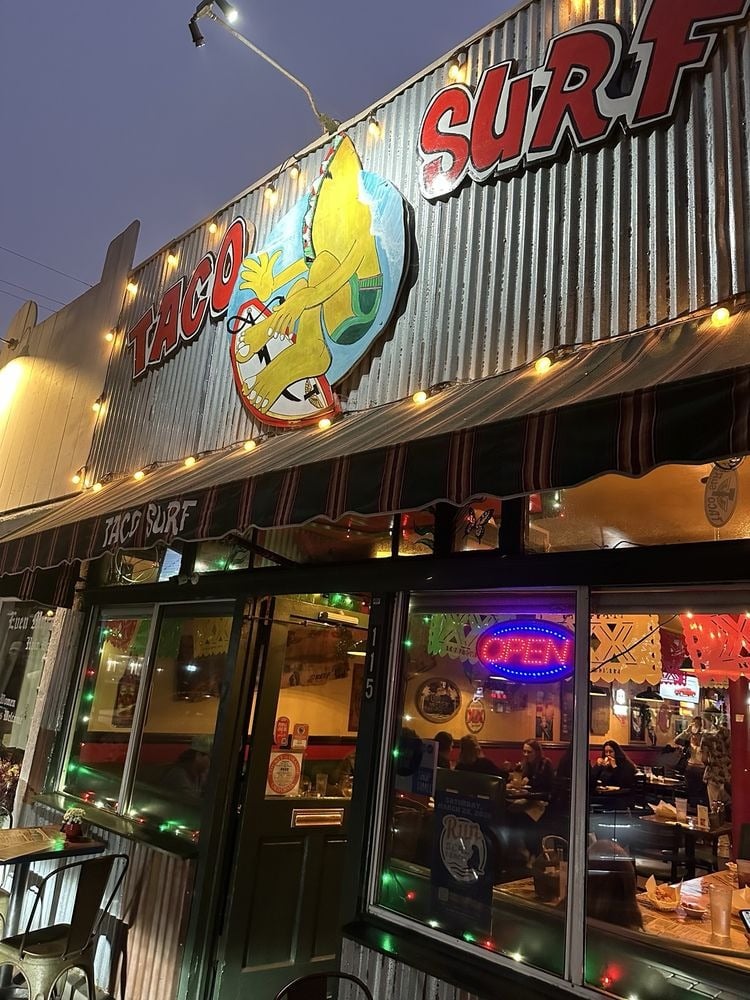 A warmly lit taco restaurant called Taco Surf, with a corrugated metal exterior, colorful surfboard sign, string lights, and neon signs in the window—one of the top breakfast burrito spots, with patrons visible dining inside.