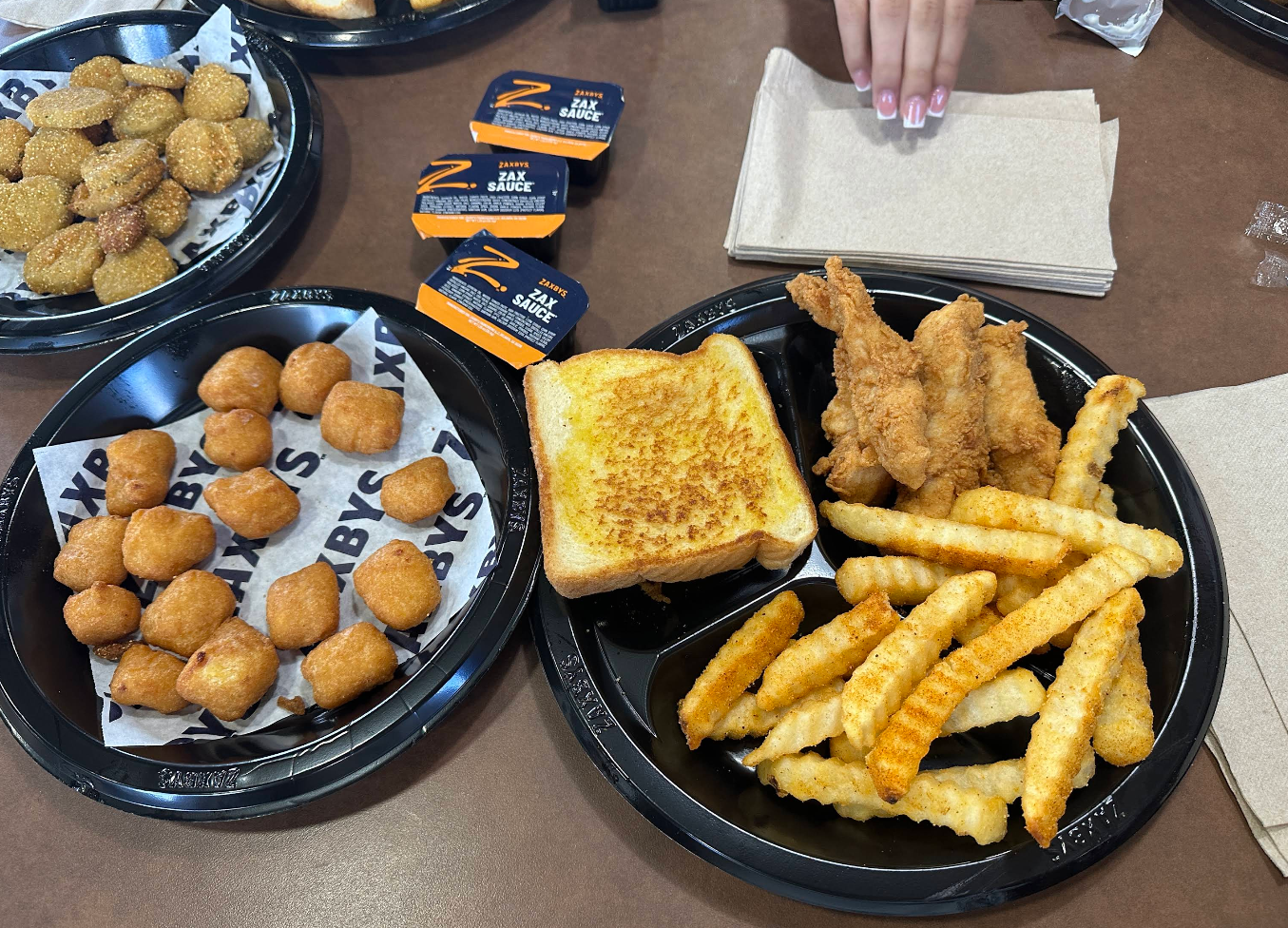A meal inspired by popular fried chicken chains: crispy chicken tenders, crinkle-cut fries, Texas toast, dipping sauces, a hand reaching for napkins, and a plate of fried cheese bites on a brown table.