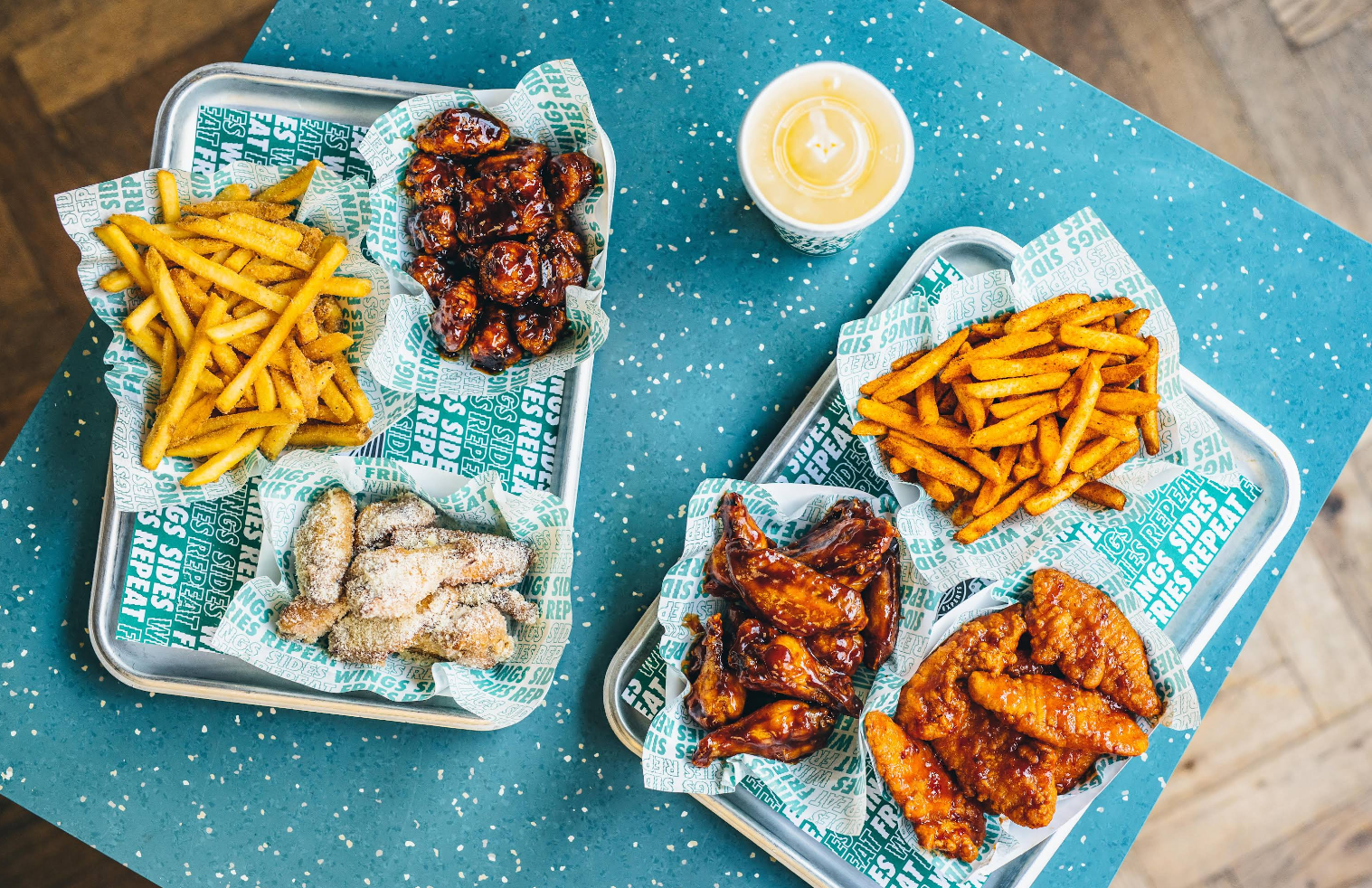 Three trays of fried food from popular fried chicken chains sit on a blue speckled table: golden fries, sauced chicken wings, breaded wings, and a cup of drink, all served on branded paper.