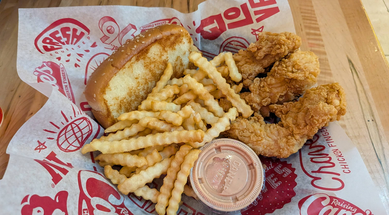 A basket from one of the top fried chicken chains, featuring three fried chicken tenders, crinkle-cut French fries, Texas toast, and dipping sauce, all served on red and white branded paper atop a wooden table.