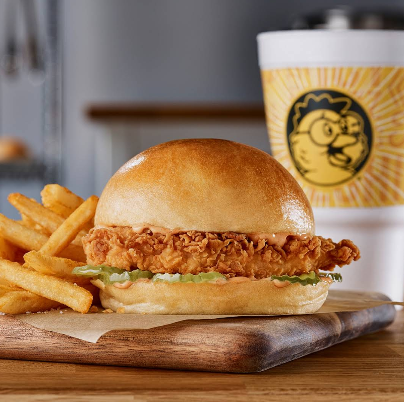 A crispy fried chicken sandwich with pickles and sauce on a shiny bun, served with golden French fries on a wooden board—just like the favorites at top fried chicken chains. A large drink cup with a chicken logo sits in the background.