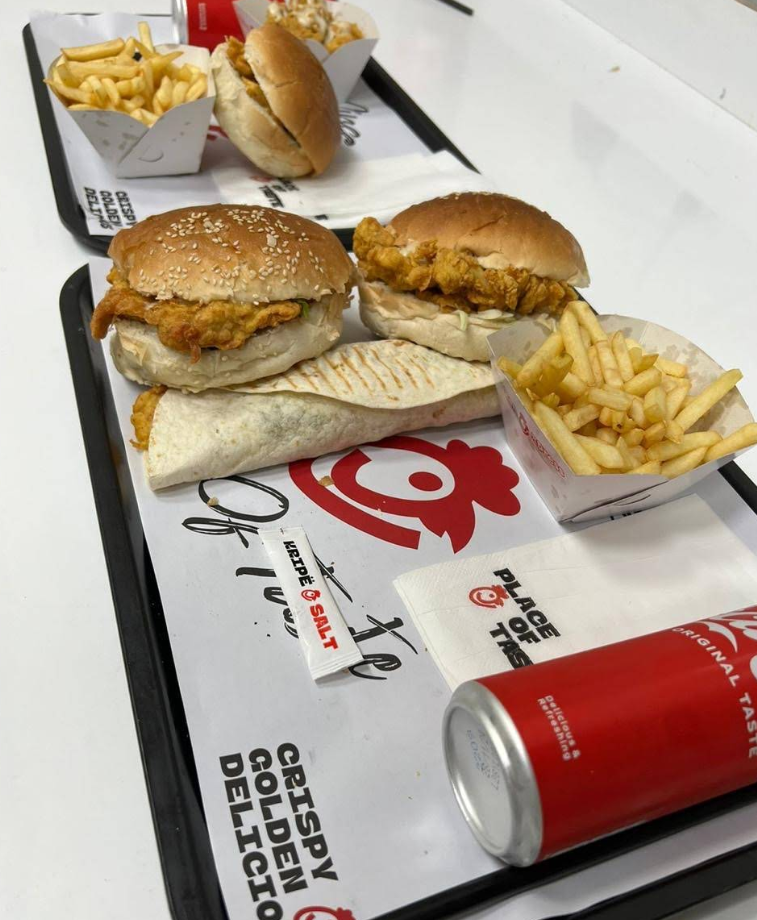 A tray with two fried chicken burgers, a wrap, French fries, and a red soda can—perfect for fans of popular fried chicken chains. Another tray with a burger and fries sits in the background on branded placemats.