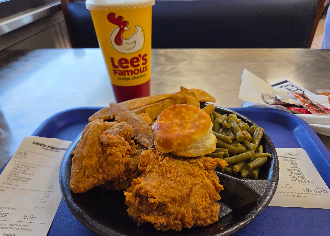 A tray from one of the top fried chicken chains features crispy chicken pieces, a biscuit, green beans, potato wedges, and a large Lee’s Famous Recipe Chicken drink. Receipts and condiment packets sit beside the tray on a restaurant table.