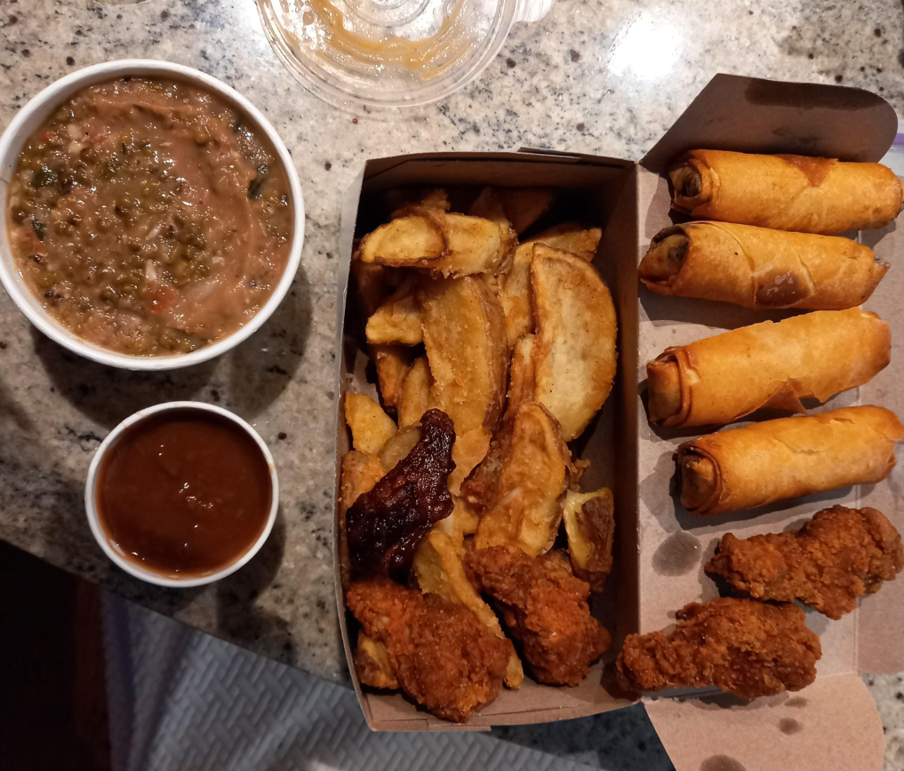 A meal inspired by fried chicken chains featuring a box of crispy chicken strips, potato wedges, and spring rolls, served with brown dipping sauce and a bowl of refried beans on a granite counter.