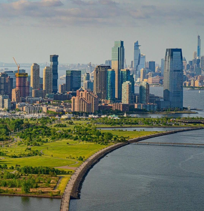 Aerial view of a curved walkway along the waterfront with a green park and modern city skyline in the background, under a partly cloudy sky—a peaceful escape from everyday worries like credit card debt.