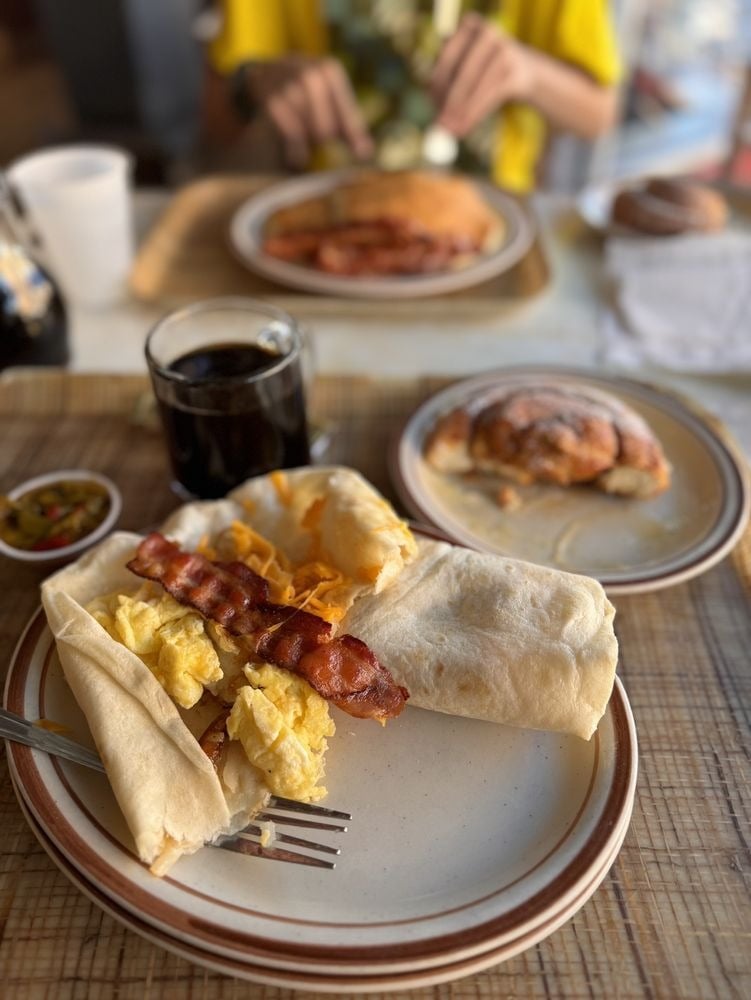 A breakfast scene with a plate featuring a wrap filled with scrambled eggs, bacon, and cheese—like those at top breakfast burrito spots—a glass of dark beverage, salsa, a cinnamon roll, and another breakfast plate in the background.