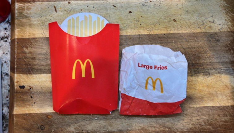 Two McDonald's fry containers on a wooden surface: one clean, empty, and upright, the other used, crumpled, and labeled "Large Fries." Both display the yellow McDonald's "M" logo.