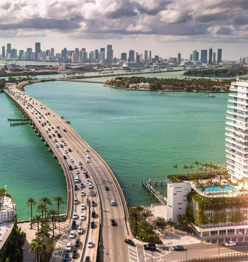 Aerial view of cars driving over a long bridge surrounded by turquoise water, with palm trees and tall buildings in the foreground—offering a striking perspective much like navigating through credit card debt toward a distant city skyline under partly cloudy skies.