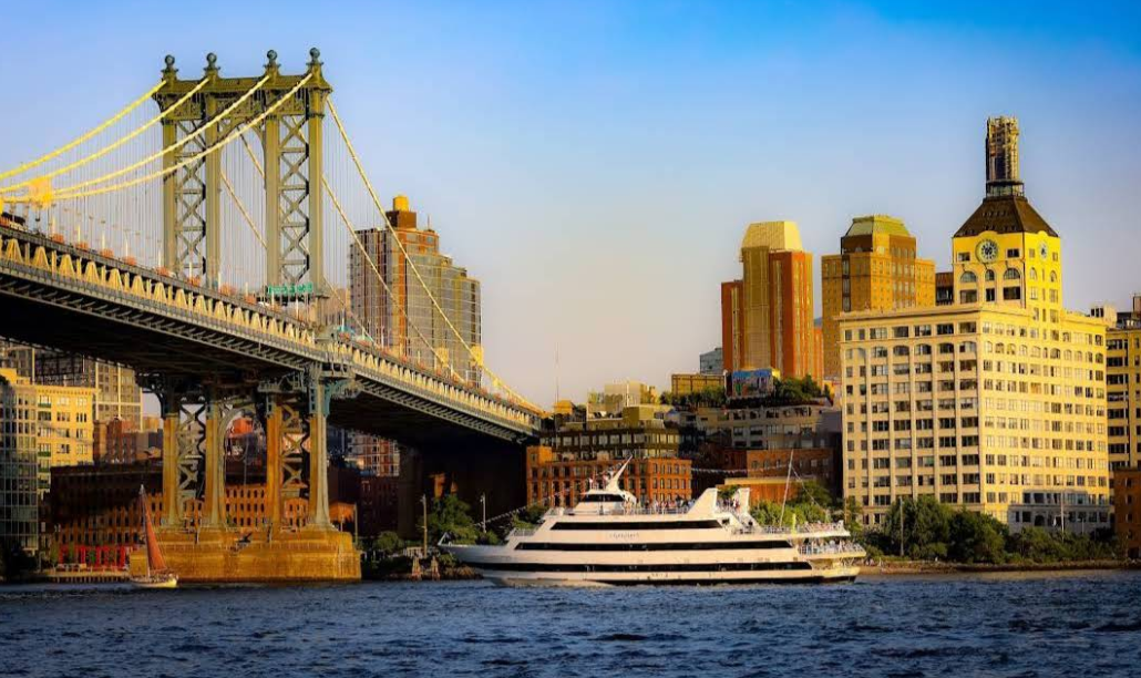 A large white yacht sails on a river beneath the Manhattan Bridge, cruising past sunlit buildings of New York City, including the iconic Domino Sugar Refinery—reminding onlookers to leave their credit card debt worries behind.