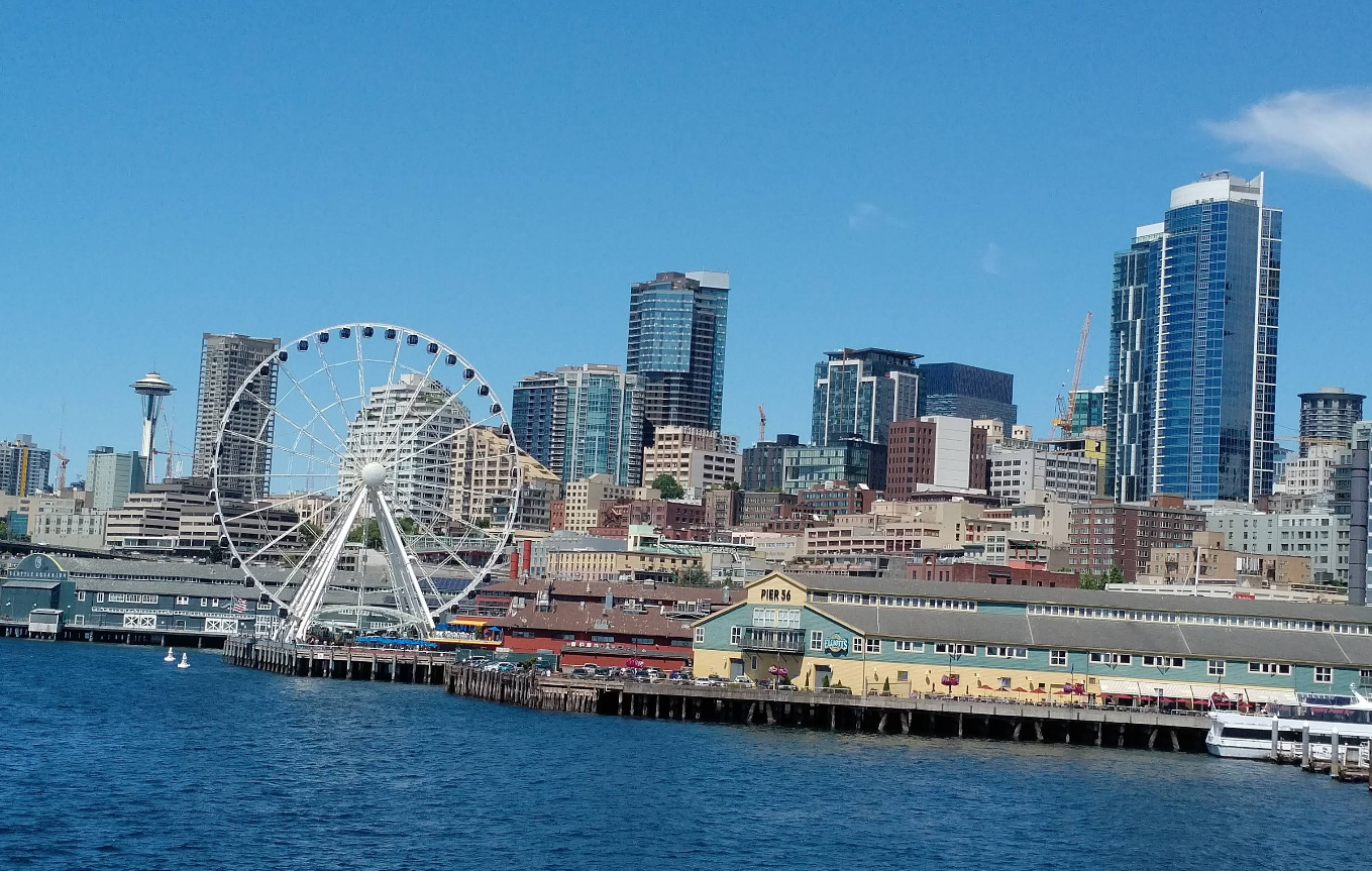 The Seattle waterfront features a large Ferris wheel, piers, downtown skyscrapers, and the Space Needle under a clear blue sky—a picture-perfect scene that might make you forget about credit card debt for a moment.