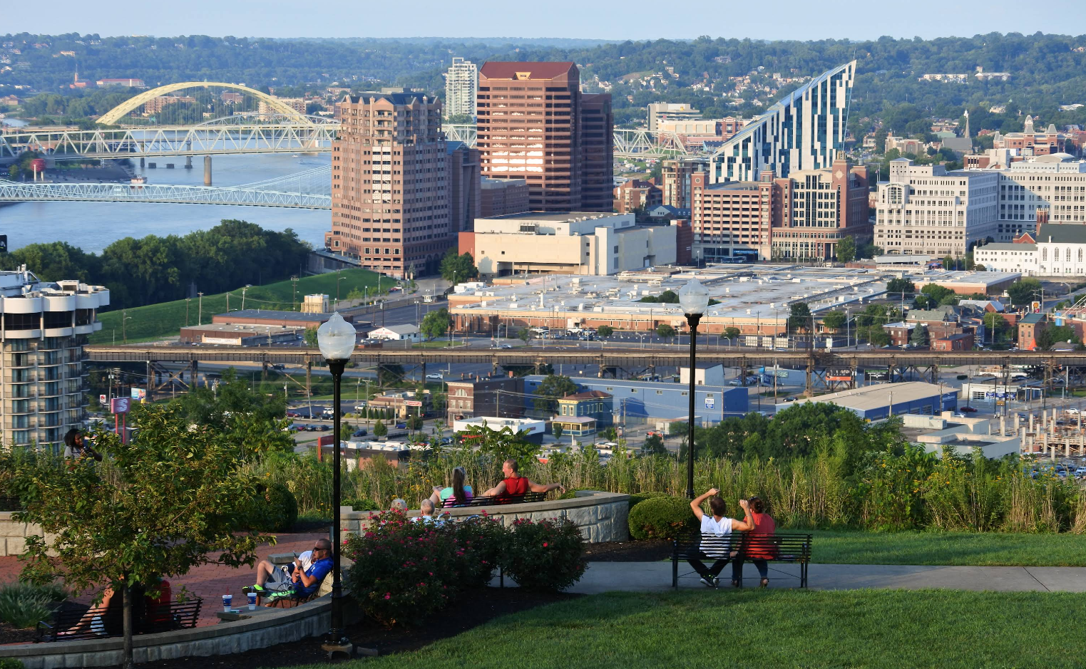 People sit on benches and relax in a park overlooking a cityscape with tall buildings, a river, and bridges in the background on a clear day, momentarily leaving worries like credit card debt behind.