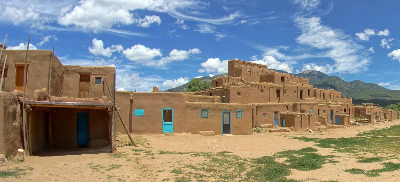 Adobe-style pueblo buildings with blue doors and windows sit under a bright blue sky, framed by green mountains and open grassy land—a peaceful escape far from the worries of credit card debt.
