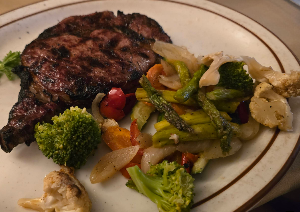 A grilled steak served on a white plate with mixed roasted vegetables—including broccoli, asparagus, cauliflower, red peppers, and onions—captures the signature flavors of a steakhouse on the West Coast.