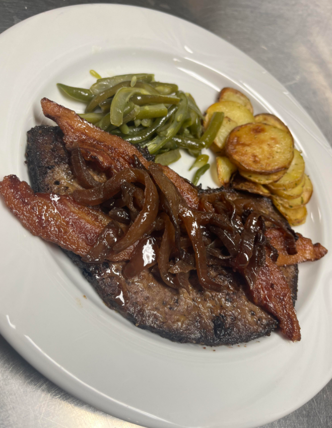 A white plate with a cooked beef steak topped with caramelized onions and two strips of bacon, served with sautéed green beans and sliced roasted potatoes—classic foods that boomers still eat and love.