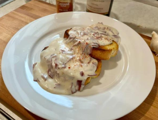 Two slices of toasted bread on a white plate topped with creamy chipped beef gravy—a classic among foods that boomers still eat—sprinkled lightly with red seasoning and set on a wooden surface.