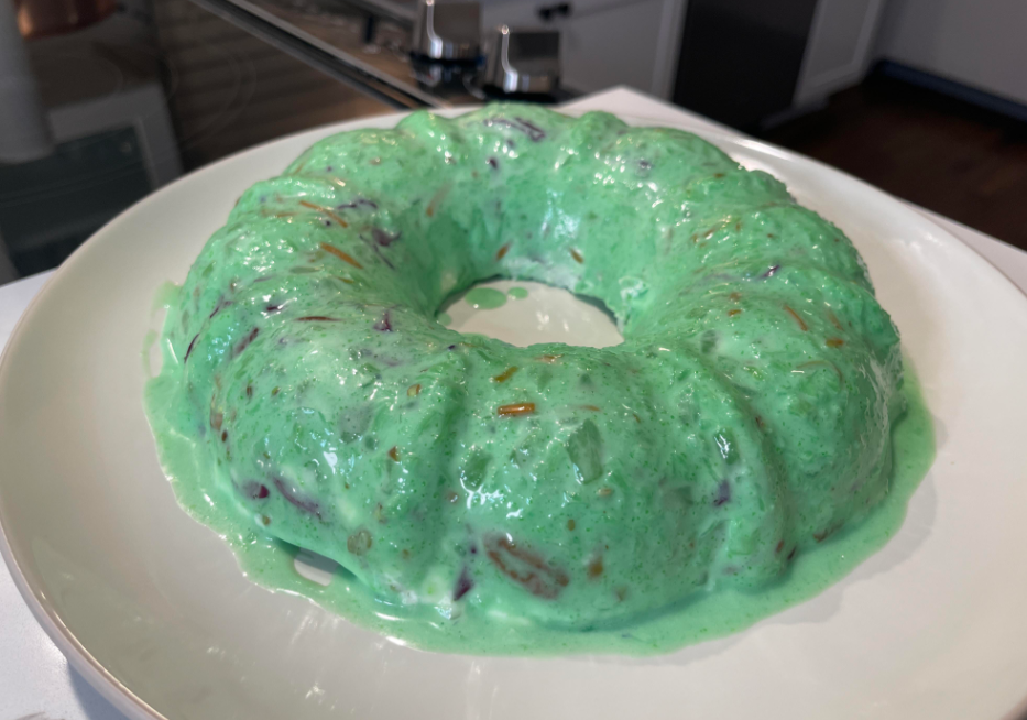 A classic from the list of foods that boomers still eat, this green, gelatin-based salad in a bundt shape sits on a white plate, with bits of fruit inside and slightly melting edges.