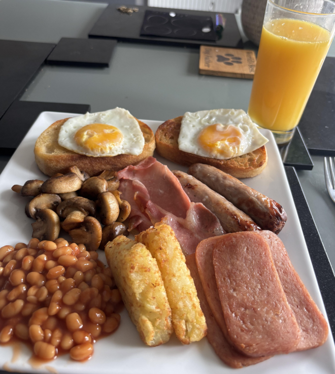 A full English breakfast on a white plate with fried eggs on toast, mushrooms, baked beans, hash browns, bacon, sausages, and sliced luncheon meat—classic foods that boomers still eat. A glass of orange juice sits beside the plate on the table.