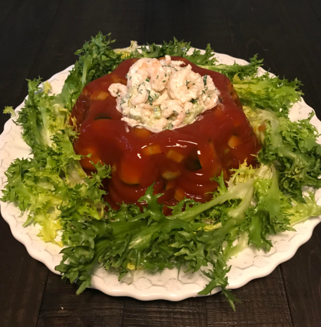 A molded tomato aspic garnished with chopped vegetables sits on a white plate, surrounded by fresh curly lettuce, and topped with a creamy shrimp salad—a classic among foods that boomers still eat.