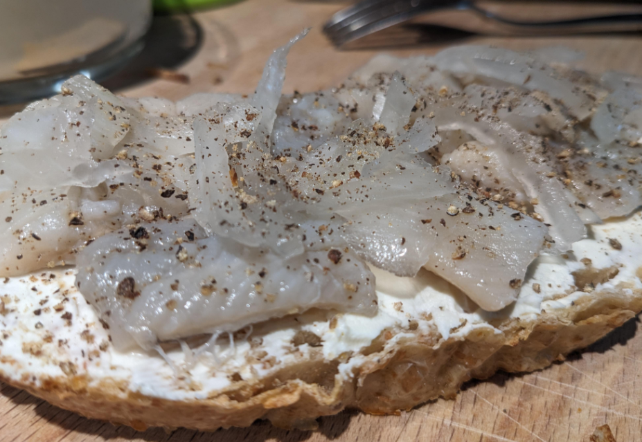 A close-up of an open-faced sandwich, a classic among foods that boomers still eat, with slices of raw fish, thin onion strips, ground black pepper, and a layer of white spread on rustic bread, placed on a wooden surface.
