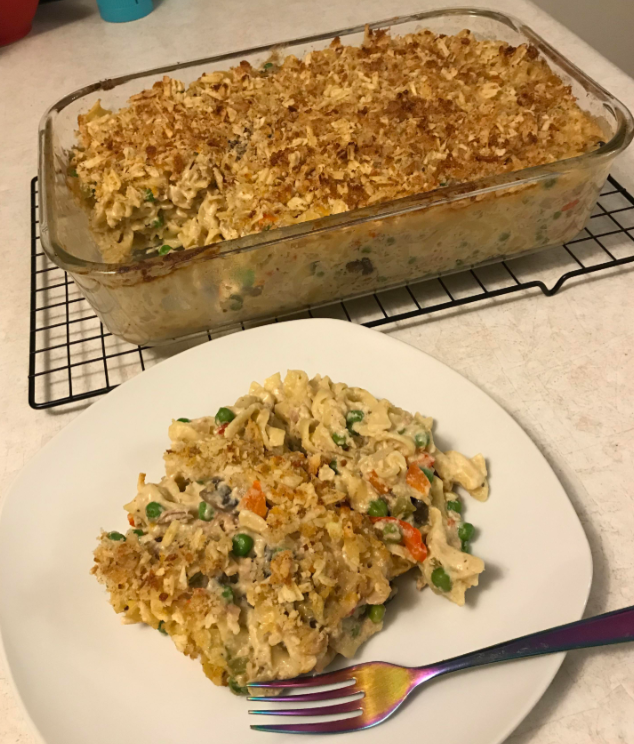 A serving of noodle casserole with peas and carrots—one of those classic foods that boomers still eat—is on a white plate with a colorful fork. The rest sits in a glass baking dish on a cooling rack in the background.