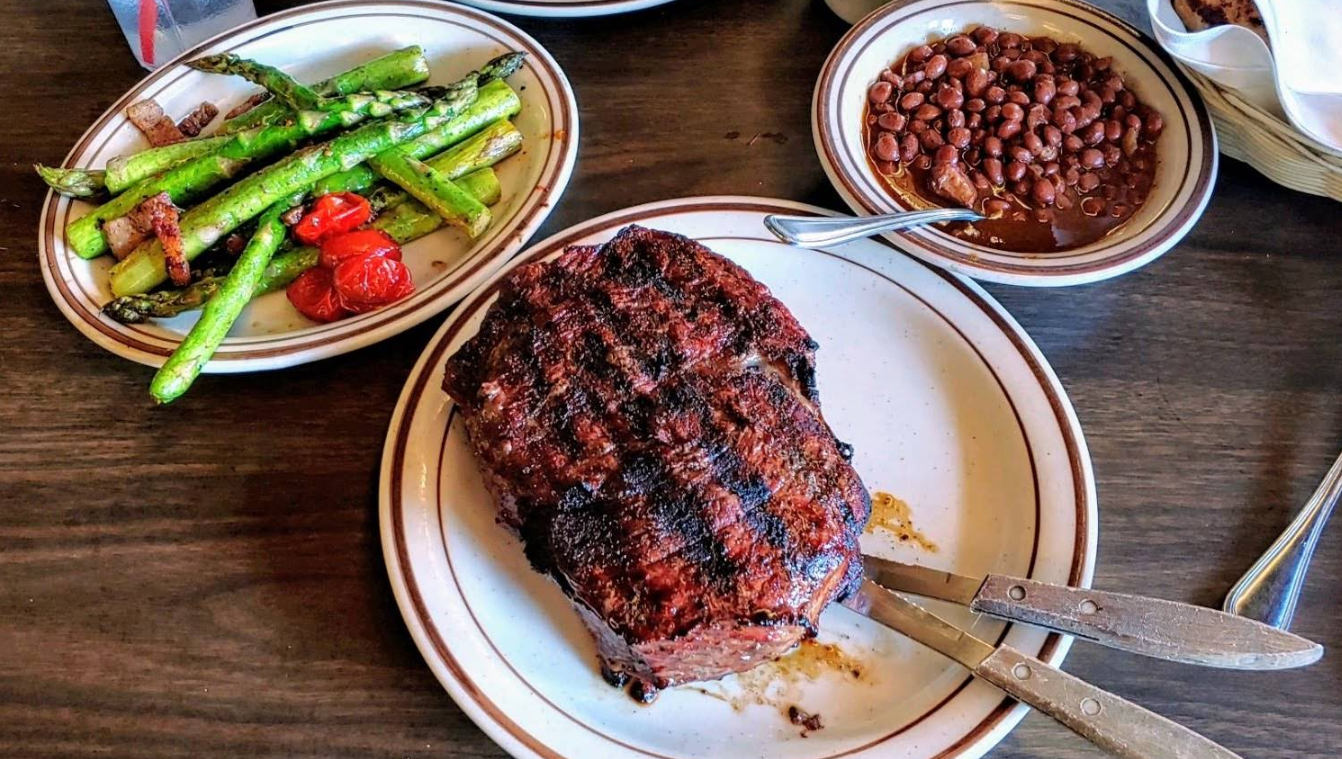 A grilled steak sits on a plate with a knife and fork, accompanied by roasted asparagus, tomatoes, and a bowl of baked beans—classic flavors you’d find at a steakhouse on the West Coast, all served on a rustic wooden table.